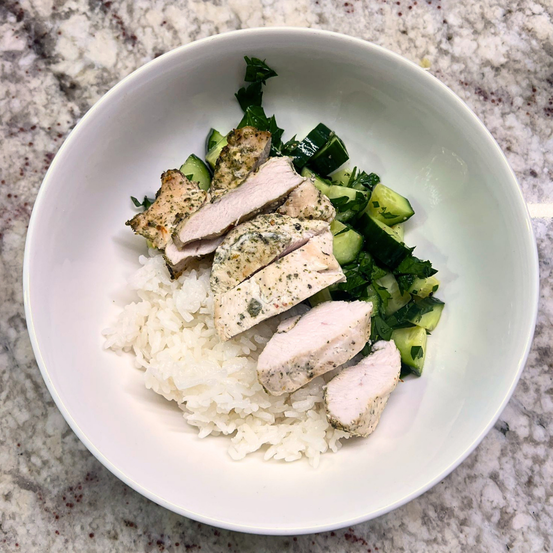 White bowl with rice, sliced chicken, and cucumber salad on a granite countertop.